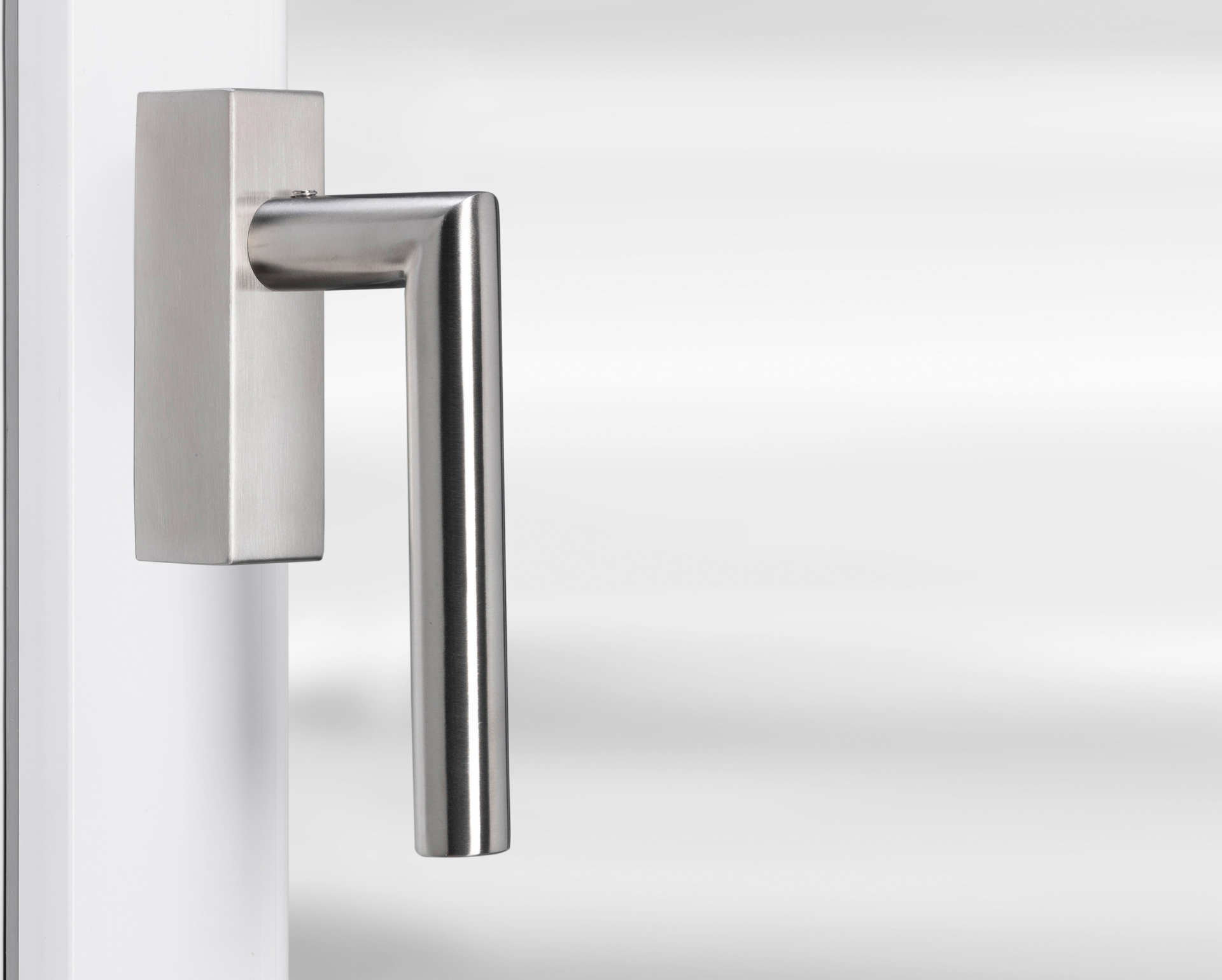 Eurolaton | Eine Nahaufnahme eines modernen, metallischen Türgriffs und Fenstergriffe aus Edelstahl, angebracht an einer weißen Tür vor einem unscharfen hellgrauen Hintergrund. Der Griff hat ein schlankes, kantiges Design.